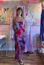 Load image into Gallery viewer, A woman modeling a long sleeveless tie-dye maxi dress with purple, pink, and red patterns, standing in a boutique with colorful painted walls and mannequins.