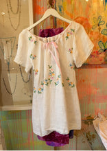 Load image into Gallery viewer, Front view of a white handmade dress tunic on a hanger, showing floral embroidery, short sleeves, and pink drawstring neckline
